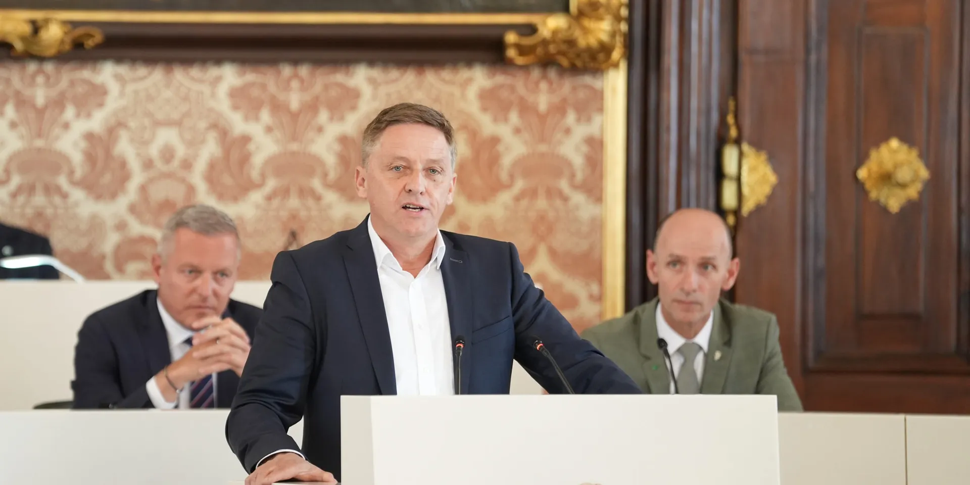 Landtagsabgeordneter Lambert Schönleitner am Rednerpult während einer Landtagssitzung. Im Hintergrund kann man Regierungsmitglieder erkennen.