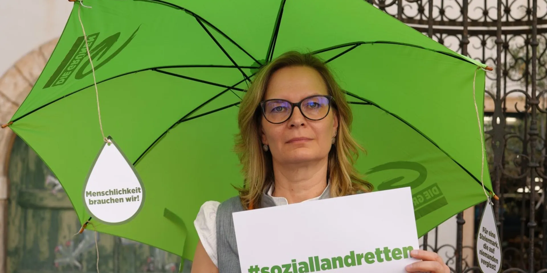 Landtagsabgeordnete Veronika Nitsche steht mit einem grünen Schirm (Rettungsschirm) und Demoschild #soziallandretten im Landhaushof