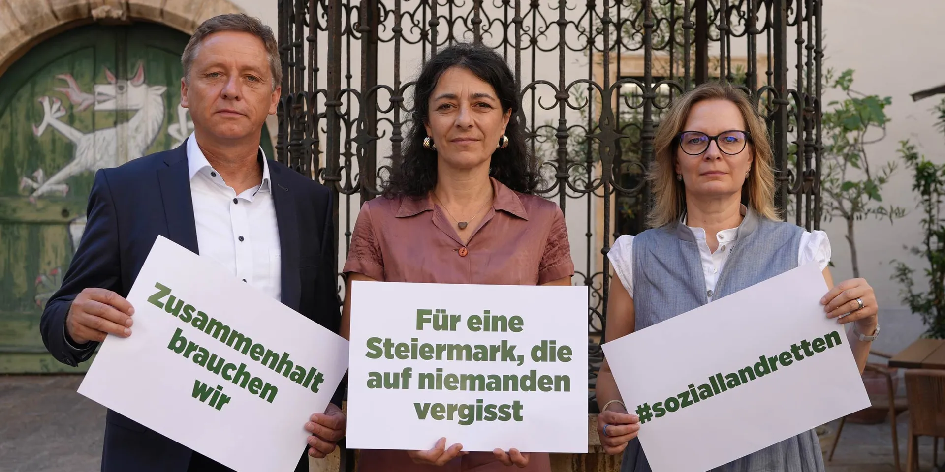 Landtagsabgeordnete Schönleitner, Krautwaschl und Nitsche stehen mit Demonstrationsschildern zu #soziallandretten im Landhaushof vor der steirischen Panther