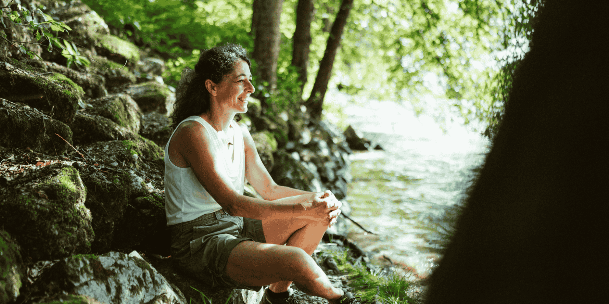 Sandra Krautwaschl, Klubobfrau der Grünen Steiermark, sitzt lächelnd auf einem moosbewachsenen Felsen am Ufer der Grenzmur und genießt die Natur.