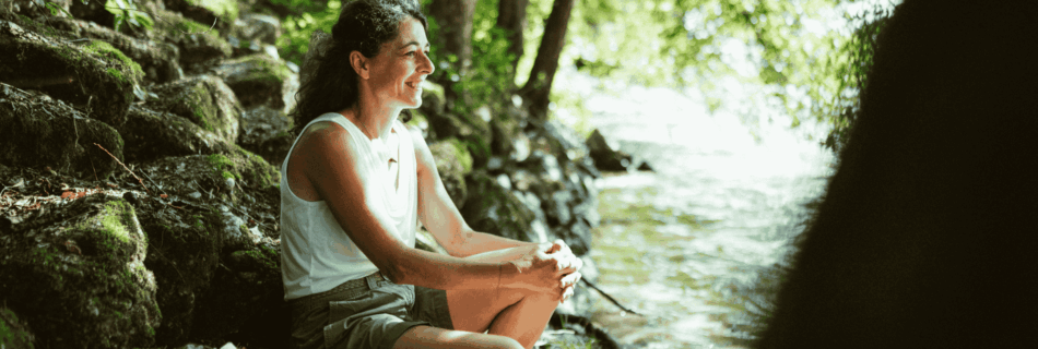 Sandra Krautwaschl, Klubobfrau der Grünen Steiermark, sitzt lächelnd auf einem moosbewachsenen Felsen am Ufer der Grenzmur und genießt die Natur.