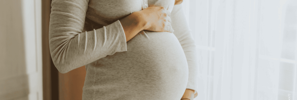 Schwangere weiblich gelesene Person steht am Fenster und hält den Schwangerschaftsbauch