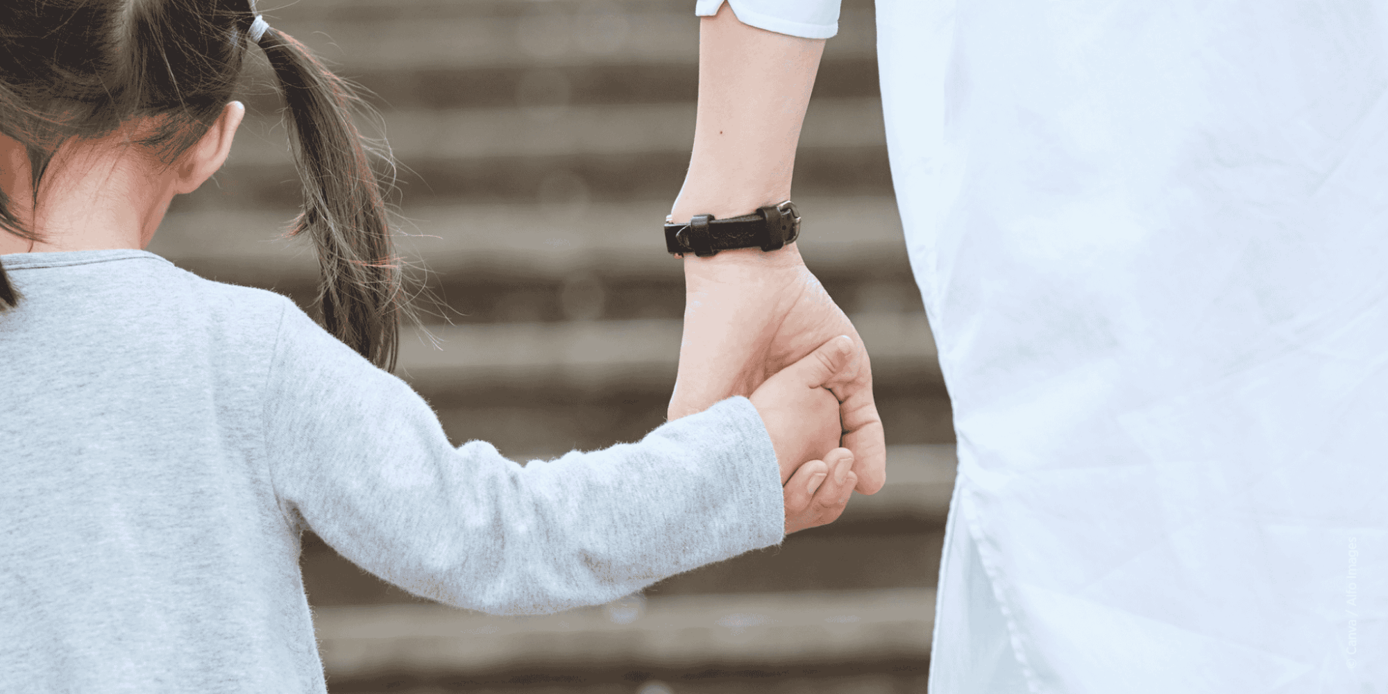 Ein Kind mit zusammengebundenem dunklen Haar, hält die Hand einer erwachsenen Person. Sie stehen vor einer Treppe.