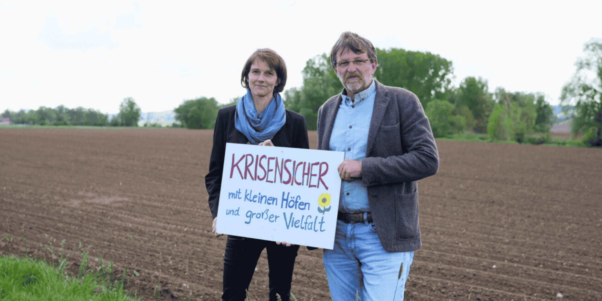 Katja Maurer und Andreas Lackner vor einem Feld mit einem Schild inder Hand: Krisensicher mit kleinen Höfen und großer Vielfalt